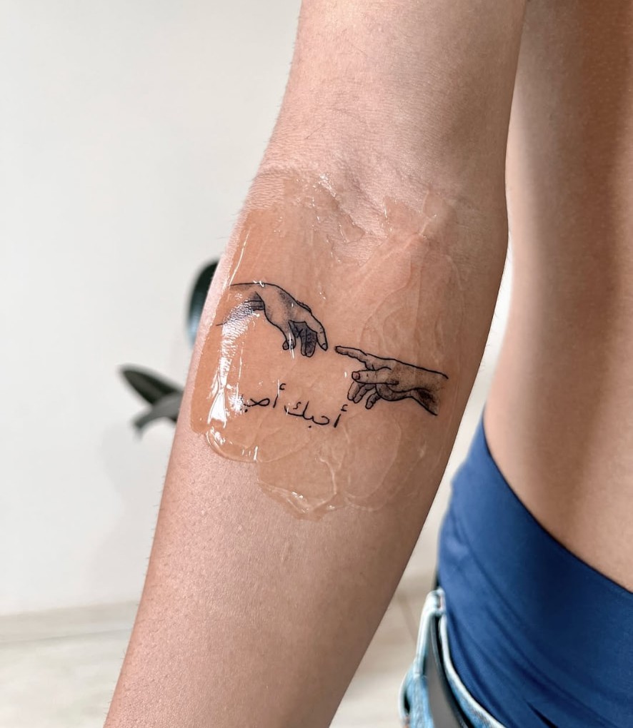 Tatuagem Criação de Adão com escrita em árabe no antebraço
